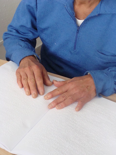Descrição de imagem: fotografia colorida de mãos tateando um livro em braille.