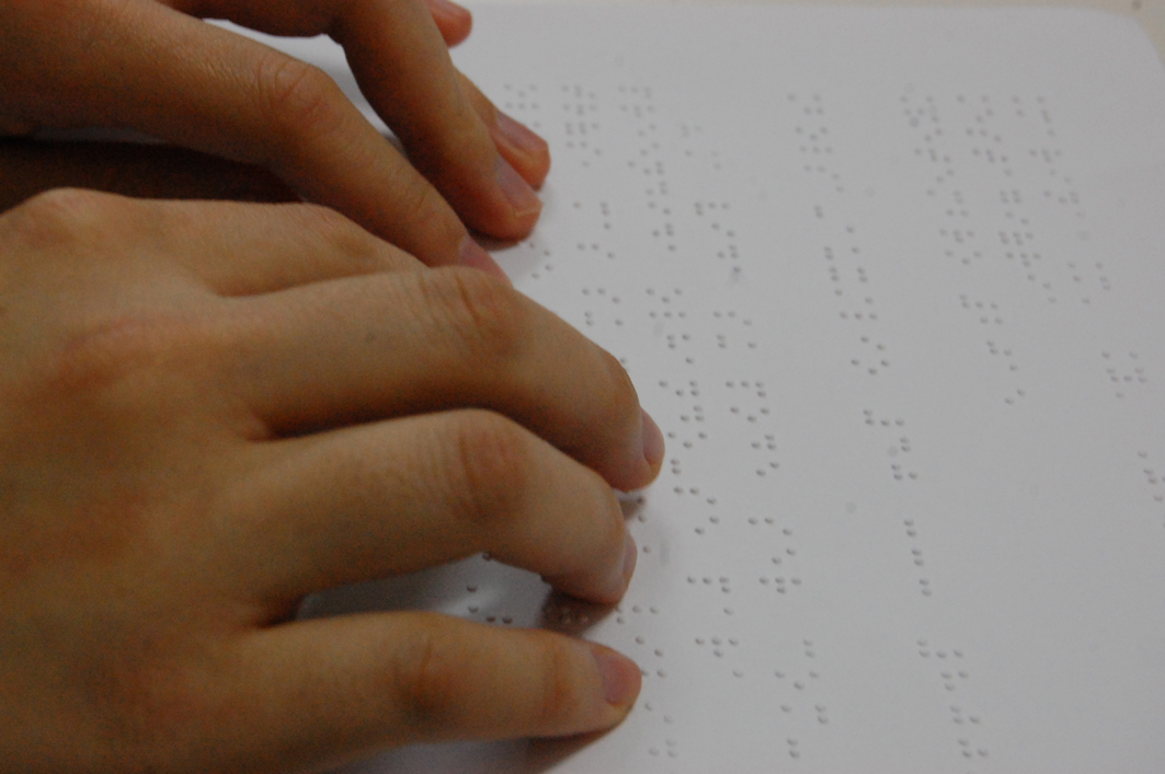 Descrição de imagem: Mãos leem um material em braille. Fim da descrição.