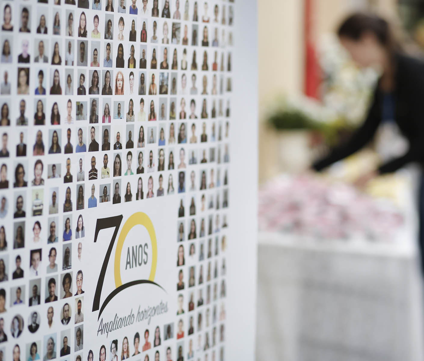 Descrição de imagem: fotografia colorida em que é possível ver um painel com várias fotos de rostos de pessoas e ao centro o logo dos 70 anos da Fundação Dorina. Ao fundo, bem desfocada, a silhueta de uma mulher. Fim da descrição.