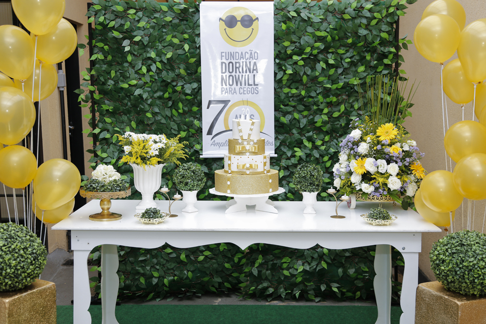 Descrição de imagem: Fotografia colorida de mesa da festa em comemoração aos 70 anos. Há um bolo de três andares, branco e dourado, cercado por vasos com folhas e flores brancas e amarelas. Fim da descrição.