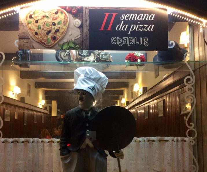 Fotografia de uma fachada de restaurante. No topo, um banner com a imagem de uma pizza em formato de coração e o texto II Semana da Pizza Chaplin. Abaixo um boneco de Charlie Chaplin com chapéu de pizzaiolo