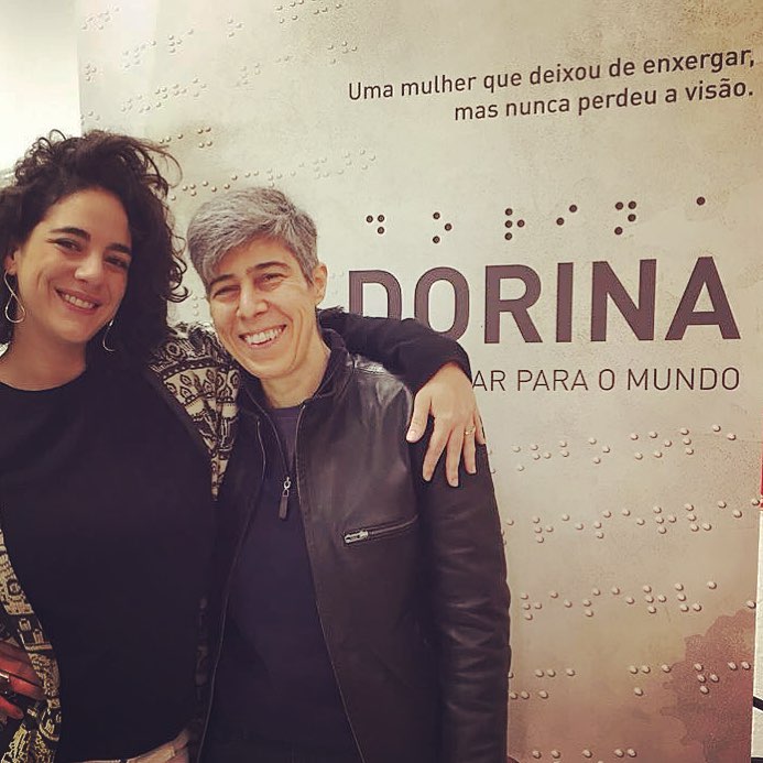 Descrição da imagem: Fotografia colorida de Marta Nowill e Lina Chamie em frente ao painel de divulgação do filme.