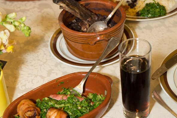 Imagem de destaque: mesa com cumbuca de feijoada, couve com torresmo e um copo de refrigerante.