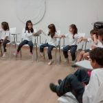 Foto de um grupo de crianças sentadas formando um semicírculo, em uma da pontas está uma responsável fazendo sinal de silêncio.