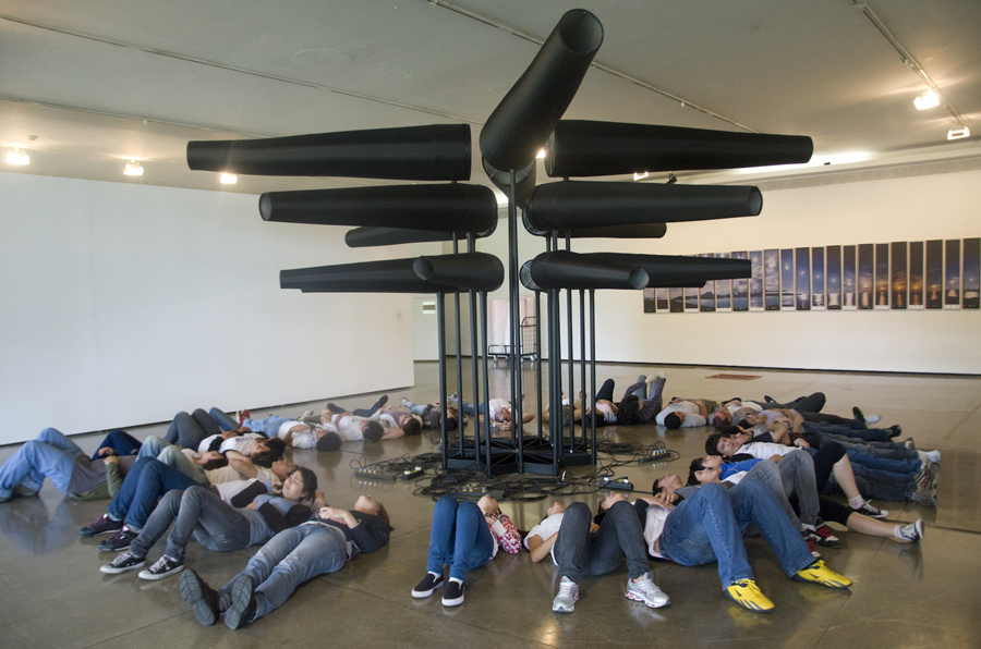 Descrição da imagem: Foto de cerca de 30 visitantes deitados no chão ao redor de uma obra no MAM. Eles formam um círculo em volta de uma estrutura metálica de cerca de 3 metros de altura com diversos tubos cônicos pretos. Fim da descrição.