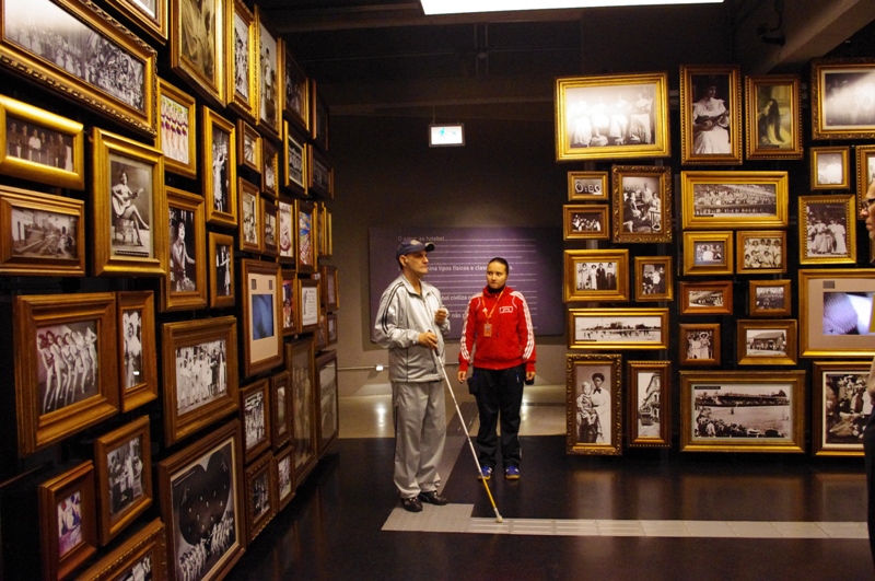 Descrição de imagem: Foto de uma sala cheia de quadros. No centro da sala duas pessoas de pé.