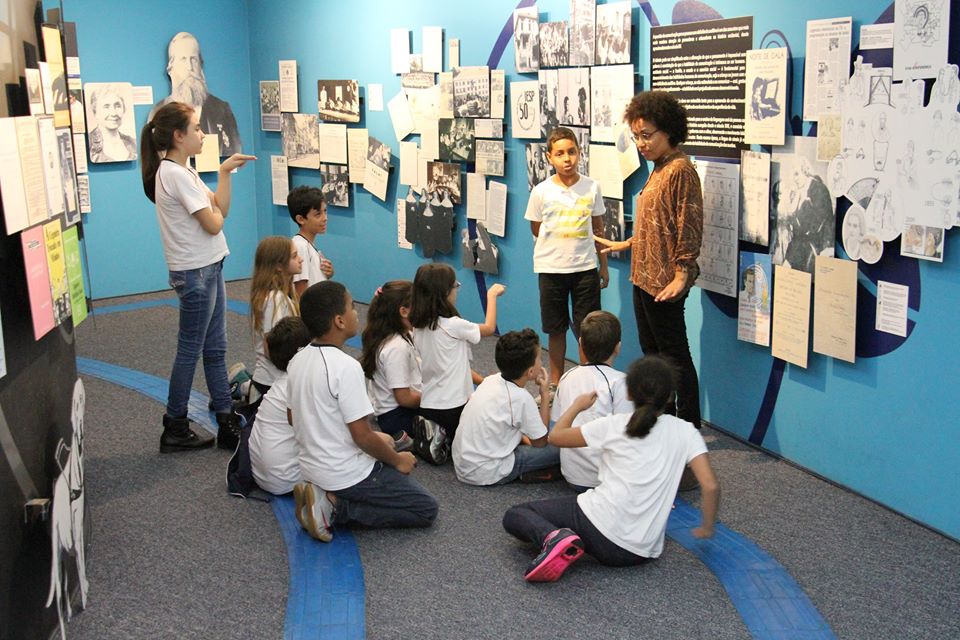 Descrição da Imagem: fotografia de um grupo de crianças em frente a um painel com diversos cartazes e figuras. A educadora está de costas para o painel. Fim da descrição.