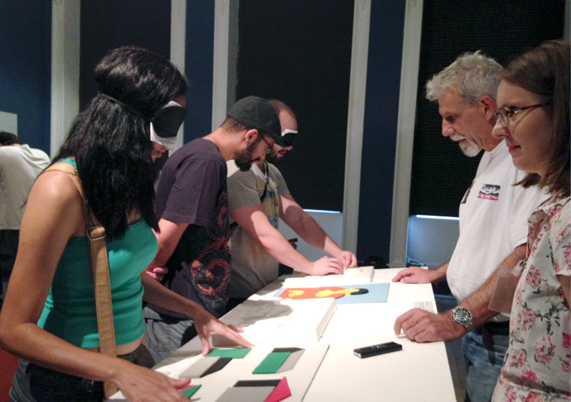 Descrição da imagem: foto de cinco pessoas ao redor de uma mesa no Museu Nacional de Belas Artes, no Rio de Janeiro. Duas delas usam vendas nos olhos e tateiam objetos geométricos em relevo na mesa.  Fim da descrição.