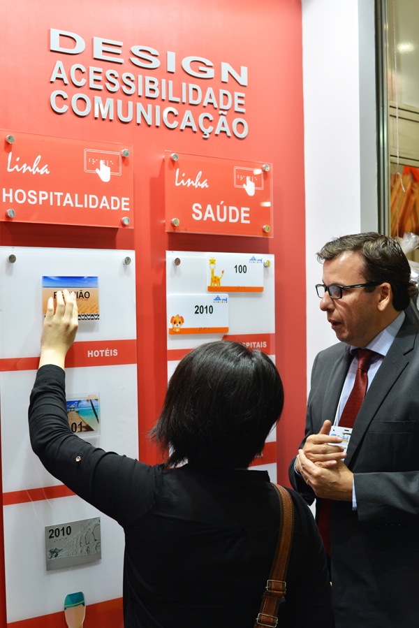 Descrição da imagem: foto de Marina tateando materiais em braille em um painel vermelho. Ela está de costas e ao seu lado está um dos expositores , que usa terno e gravata. Fim da Descrição.