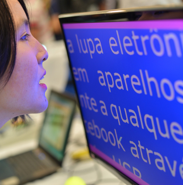 Descrição da imagem: foto de Marina lendo um texto em  um monitor com fonte ampliada . Ela está de perfil, com o rosto bem próximo à tela. Fim da descrição.