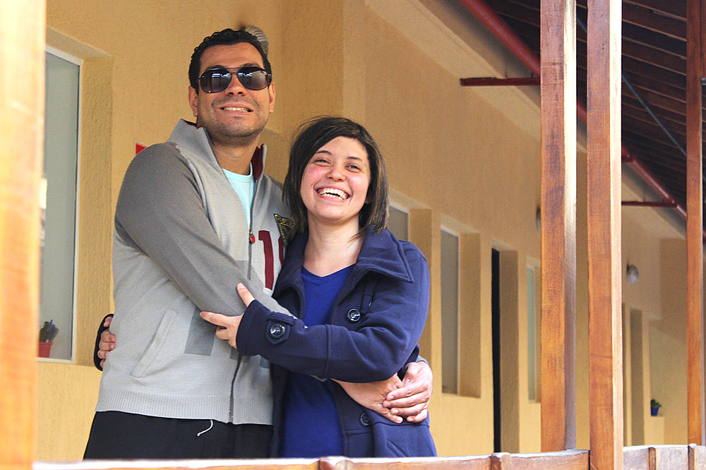 Descrição da imagem: foto de Felipe e Juliana numa área externa da Fundação Dorina. Eles estão abraçados, encostados em um parapeito de madeira, olhando para frente e sorrindo. Felipe é moreno, tem cabelos curtos e veste uma blusa de moleton cinza. Juliana tem a pele clara, cabelos lisos na altura dos ombros e usa um casaco azul marinho. Fim da descrição.