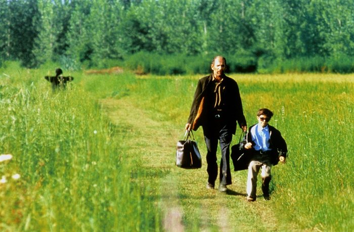 Descrição da imagem: cena do filme A cor do paraíso. Um homem e um menino caminham por uma estrada verdejante. Eles estão de frente. O homem carrega duas malas. O menino usa óculos escuros e caminha ao seu lado. Fim da descrição.