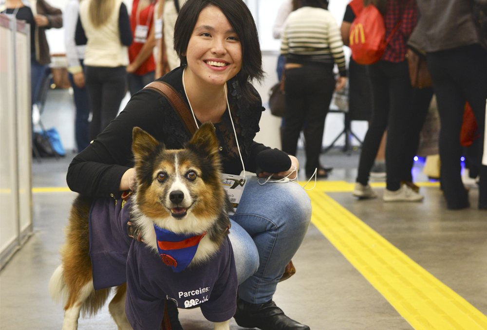 Descrição da imagem: foto colorida de Marina Yonashiro, da Fundação Dorina, abaixada ao lado de um cão terapêutico, da raça Corgi.  Marina tem traços orientais e cabelos curtos, olha pra frente e sorri. Fim da descrição.