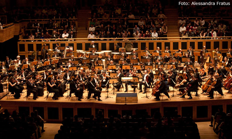 Descrição da imagem: foto da Orquestra Sinfônica do Estado de São Paulo. Dezenas de músicos estão tocando em um palco de madeira. Eles usam ternos pretos. Ao fundo, em segundo plano, está a plateia, com dezenas de pessoas sentadas. Fim da descrição.