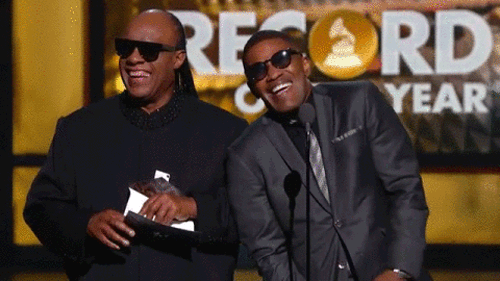 Descrição da imagem: GIF animado exibe o cantor Stevie Wonder tateando um papel em branco ao lado do ator Jamie Foxx. Ambos usam terno e óculos escuros e estão sorrindo. À frente deles há um microfone.