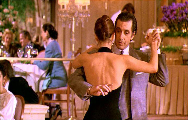 Descrição da imagem: cena do filme Perfume de Mulher. Um casal é retratado da cintura pra cima, dançando em um restaurante. Fim da descrição.