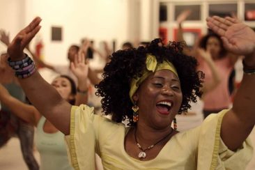 Descrição da imagem: foto de uma mulher dançando. Ela é retratada do peito pra cima, sorrindo, com os braços levantados e as palmas das mãos voltadas pra cima. A mulher é negra, usa um colar de concha, brincos, pulseiras, um lenço amarelo no cabelo e uma bata amarela. Ao fundo há diversas outras pessoas fazendo o mesmo movimento. Fim da descrição.