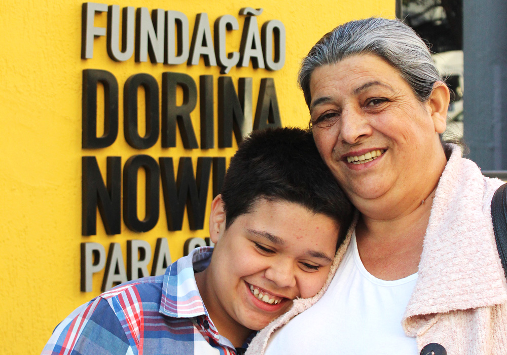 Descrição da imagem: foto de Jhonata e sua mãe Ana Lúcia. Jhonata está sorrindo com a cabeça encostada no ombro da mãe, uma senhora de pele clara e cabelos grisalhos. Ela olha pra frente e sorri. Eles estão retratados do peito pra cima, na frente de uma parede amarela com um letreiro "Fundação Dorina Nowill para Cegos"