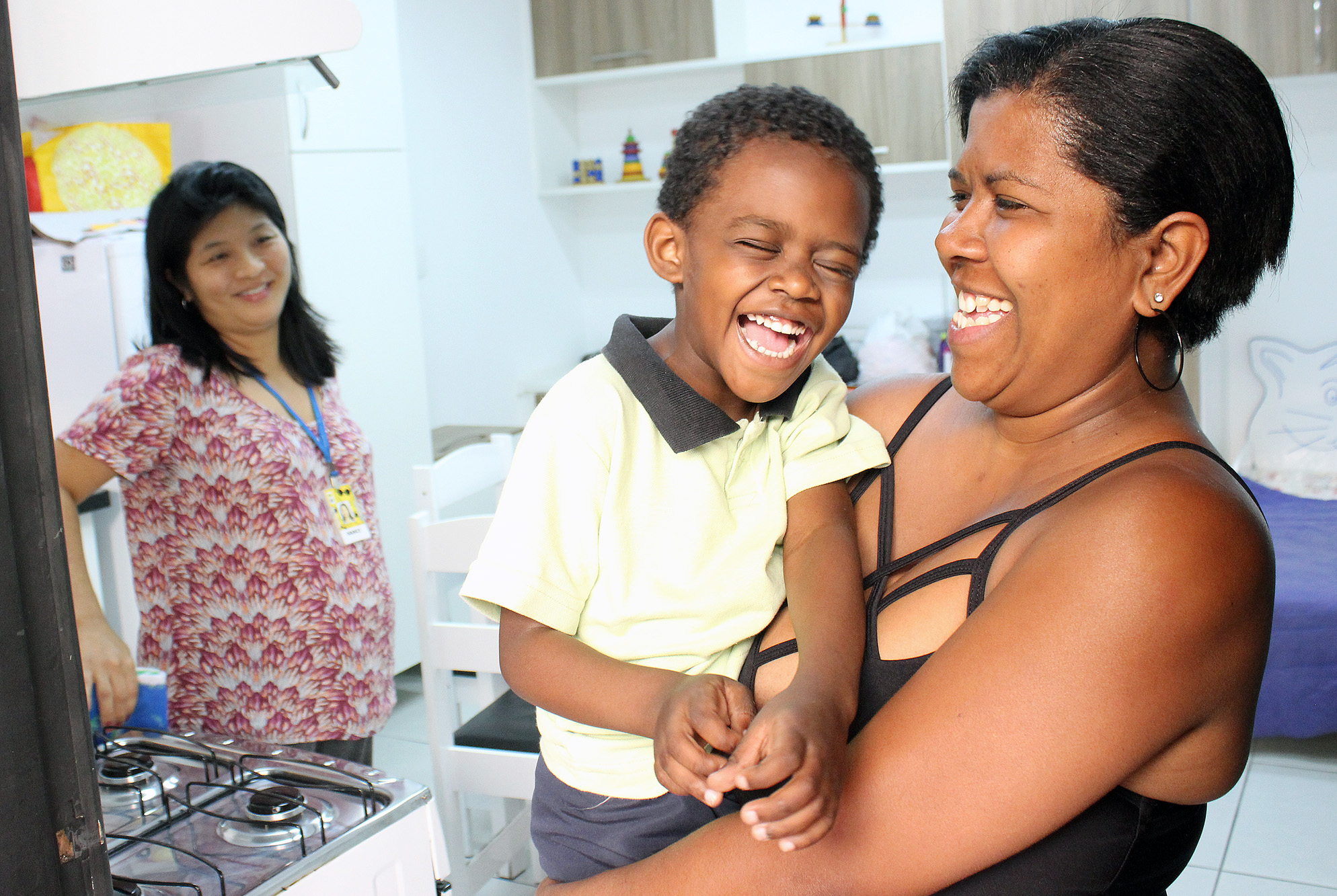 Descrição da imagem: foto de Cris no colo de sua mãe, Cristiane. Ambos são retratados da cintura pra cima e exibem um largo sorriso com os olhos semicerrados. Ao fundo, em segundo plano, está a terapeuta Nanci Hara, que observa os dois e também sorri. Eles estão em uma sala da Fundação Dorina equipada com aparelhos e móveis domésticos, como fogão, geladeira, armários e mesa de cozinha. Fim da descrição.