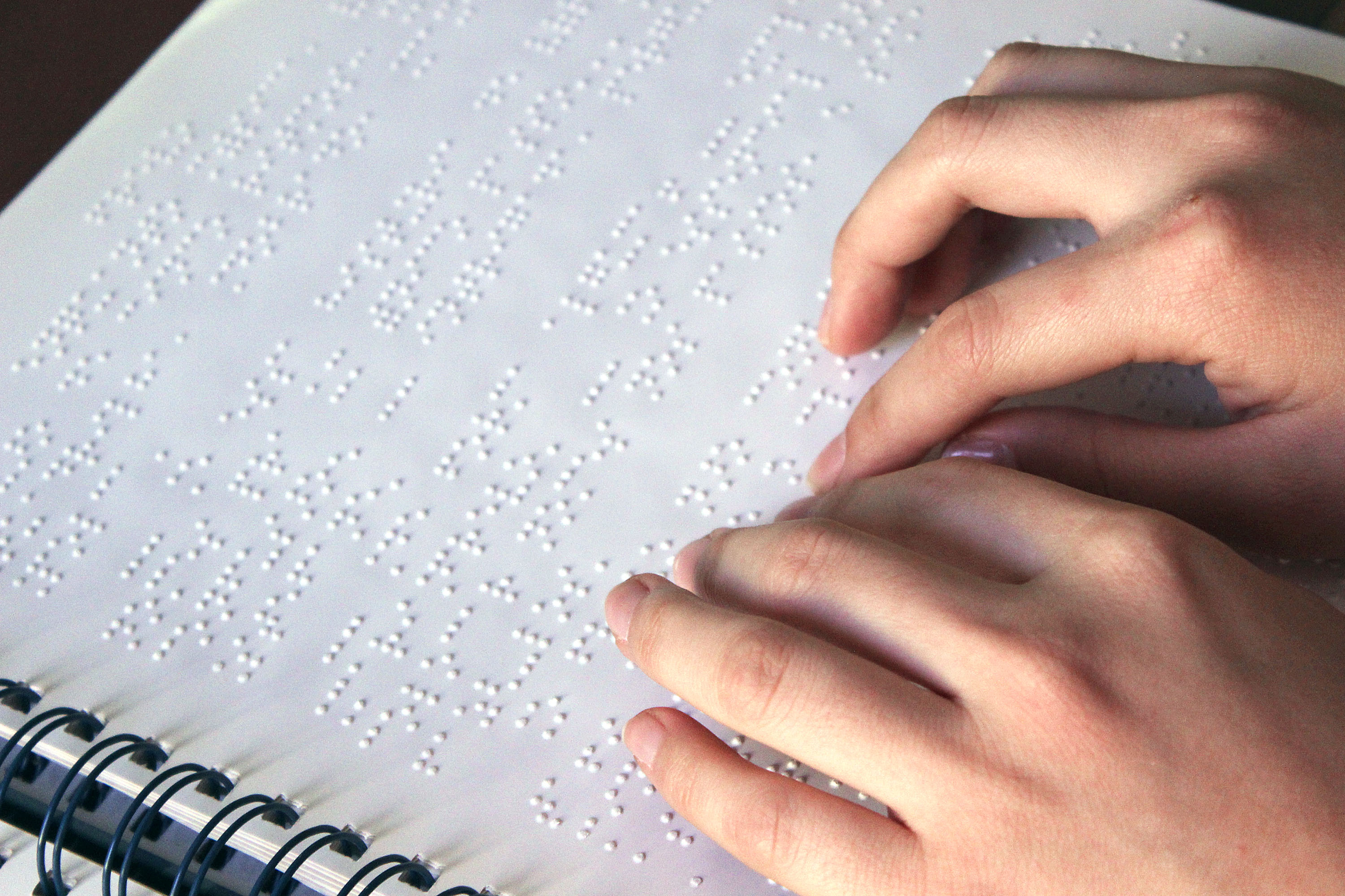 Descrição da imagem: foto de duas mãos tateando um texto em braille em uma folha branca. Fim da descrição.