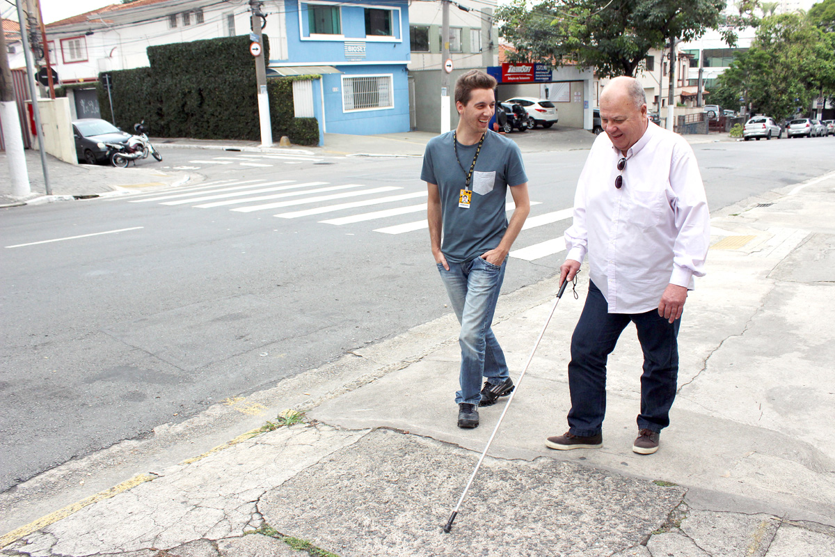 Descrição da imagem: foto de Luiz e Fabrizzio caminhando por uma calçada. Eles estão sorrindo. Luiz usa bengala e olha em direção ao chão. Ao lado esquerdo há uma rua com alguns carros estacionados e uma faixa de pedestres. Fim da descrição.