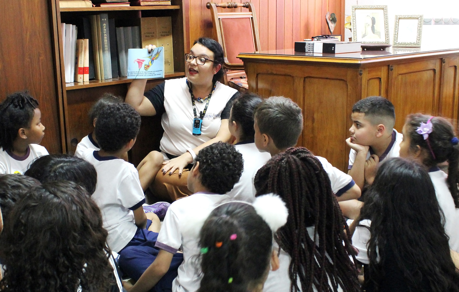 Foto de um grupo de crianças sentadas no chão junto com uma educadora da Fundação Dorina, que exibe um livro.