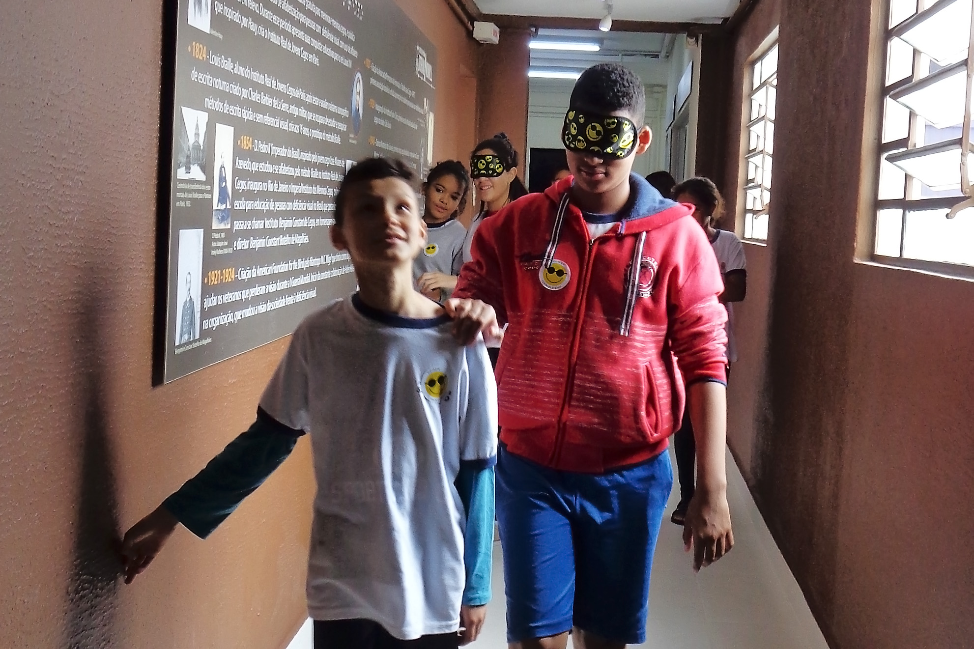 Foto de dois meninos em um dos corredores da Fundação Dorina. Um deles está vendado e com a mão no ombro do colega. Atrás deles há um grupo de crianças.