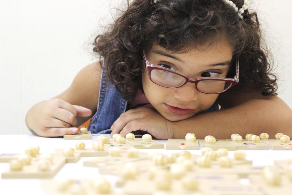 Descrição da imagem: foto de Ana Carolina, uma criança de 6 anos, manuseando peças de um alfabeto braile de madeira sobre uma mesa. Fim da descrição.