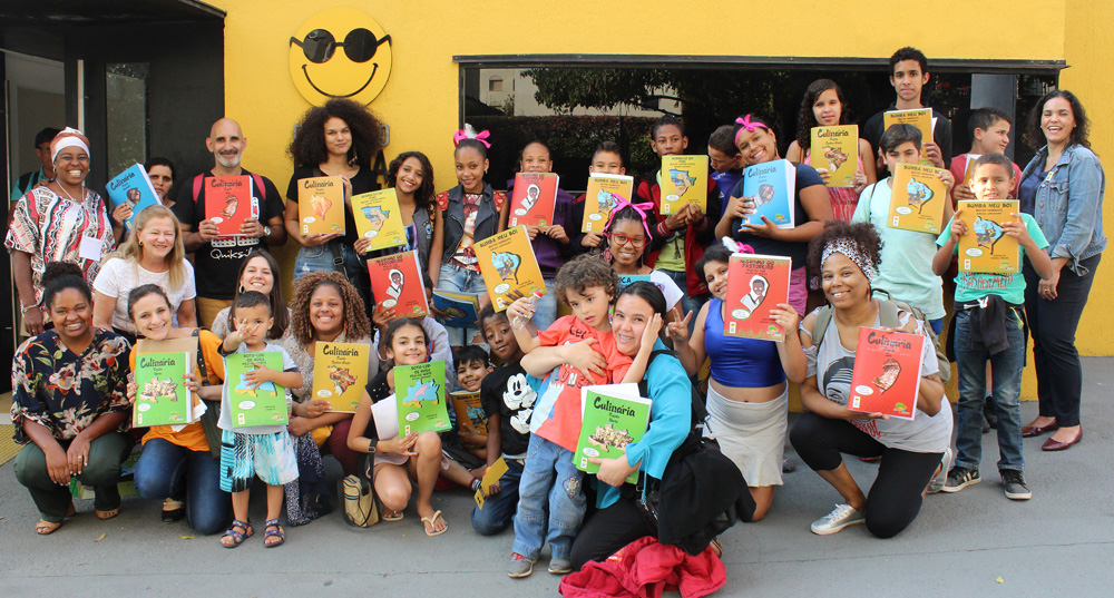 Descrição da imagem: foto de um grupo de crianças e adultos exibindo livros da Coleção Regionais. Eles olham pra frente e sorriem. Fim da descrição.