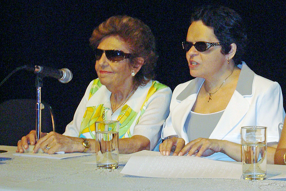 Foto de Dorina Nowill e Regina Oliveira. Elas estão sentadas com as mãos sobre a mesa, que tem um microfone e dois copos d'água. Regina está tateando um texto em braile. Ambas usam óculos escuros.