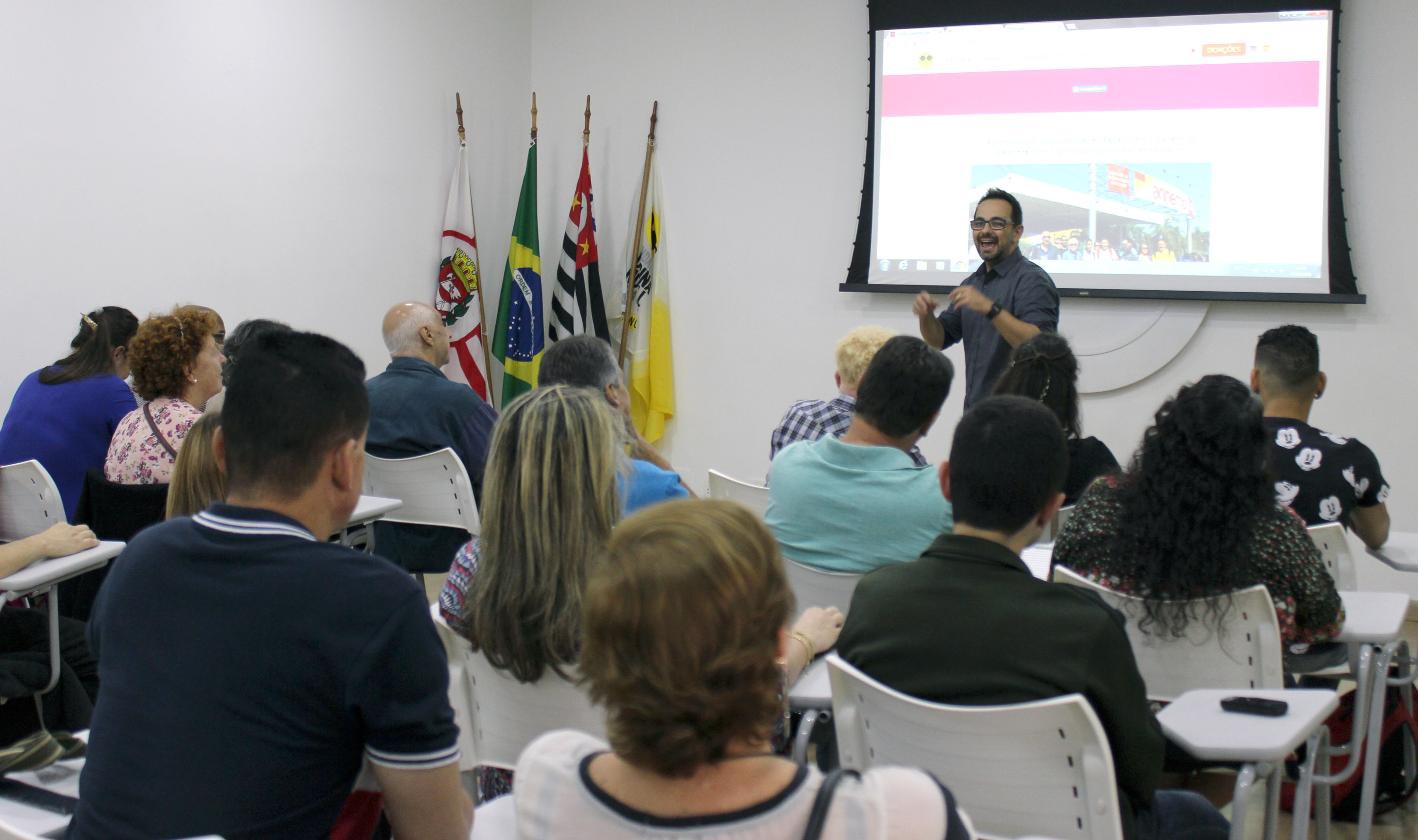 Descrição da imagem: foto de Renato Ribeiro falando para cerca de 30 pessoas no auditório da Fundação Dorina. Ele é moreno, tem cabelos curtos, veste camisa cinza, usa óculos e relógio preto. Atrás dele há um telão.