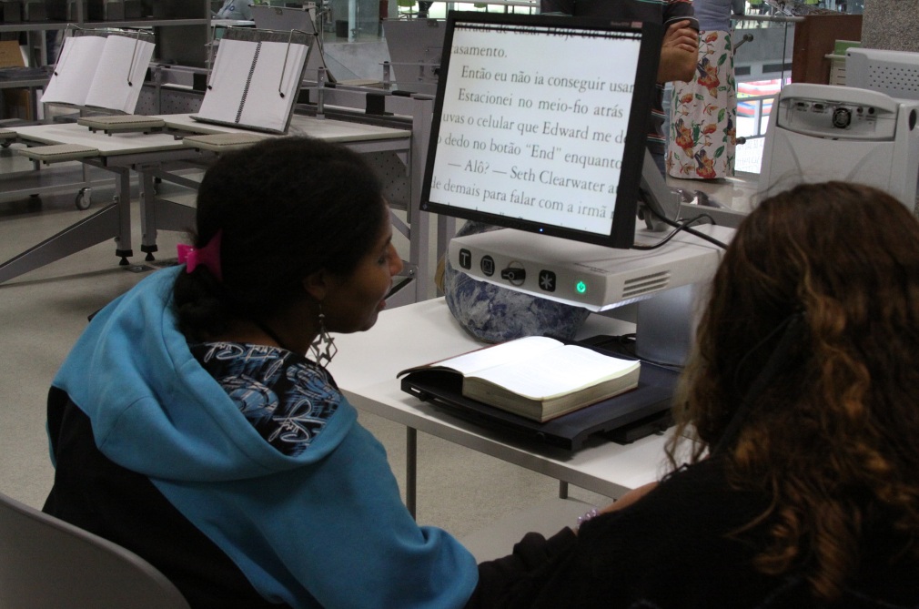 Descrição da imagem: foto de duas moças à frente de um monitor com lente de aumento digital, que exibe a página de um livro em papel. Ao fundo há mesas vazias da Biblioteca de São Paulo.