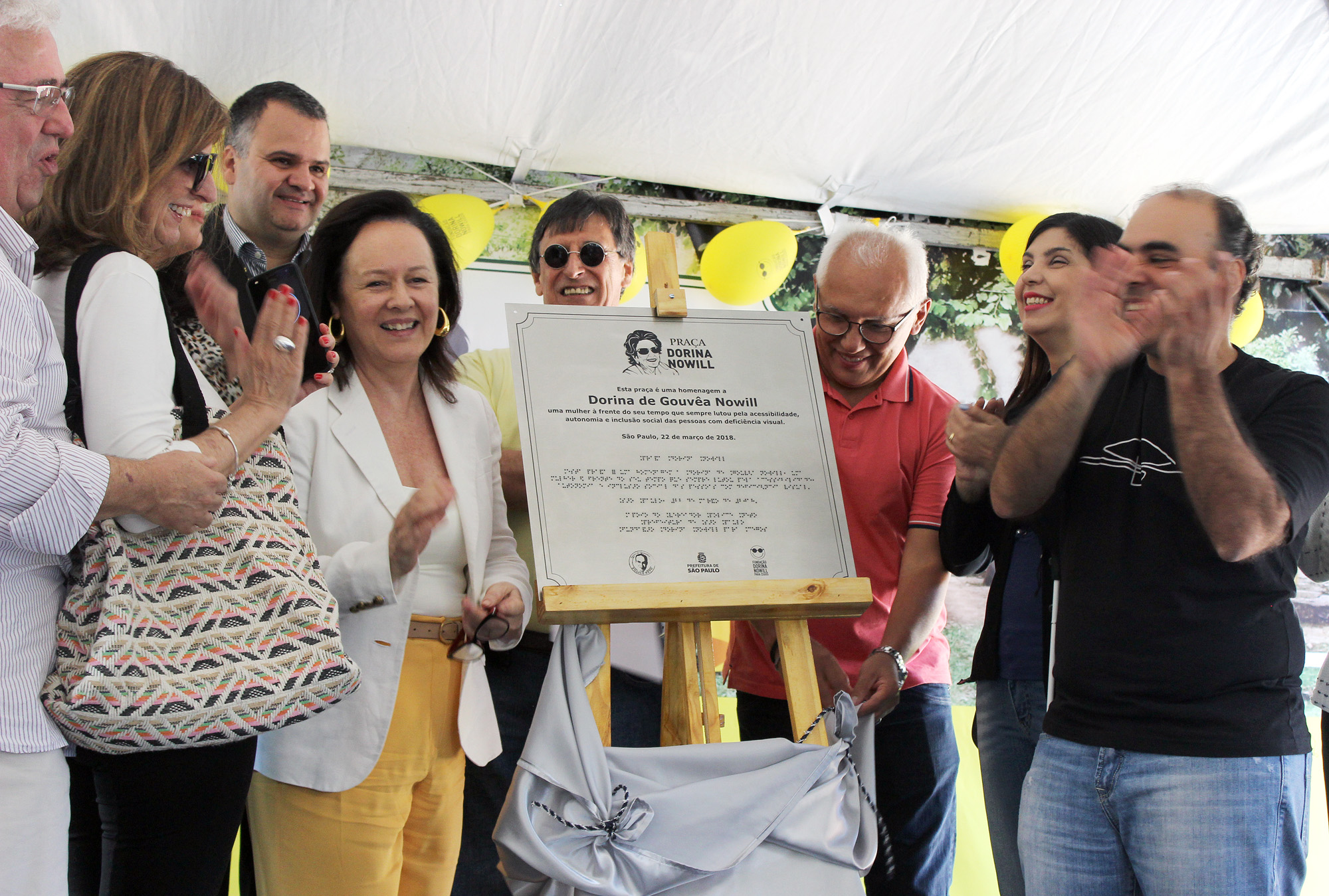 Descrição da imagem: foto de um grupo de nove pessoas em volta da placa em homenagem à Praça Dorina Nowill em um cavalete de madeira. Eles estão sorrindo e aplaudindo. Ao fundo há alguns balões amarelos.