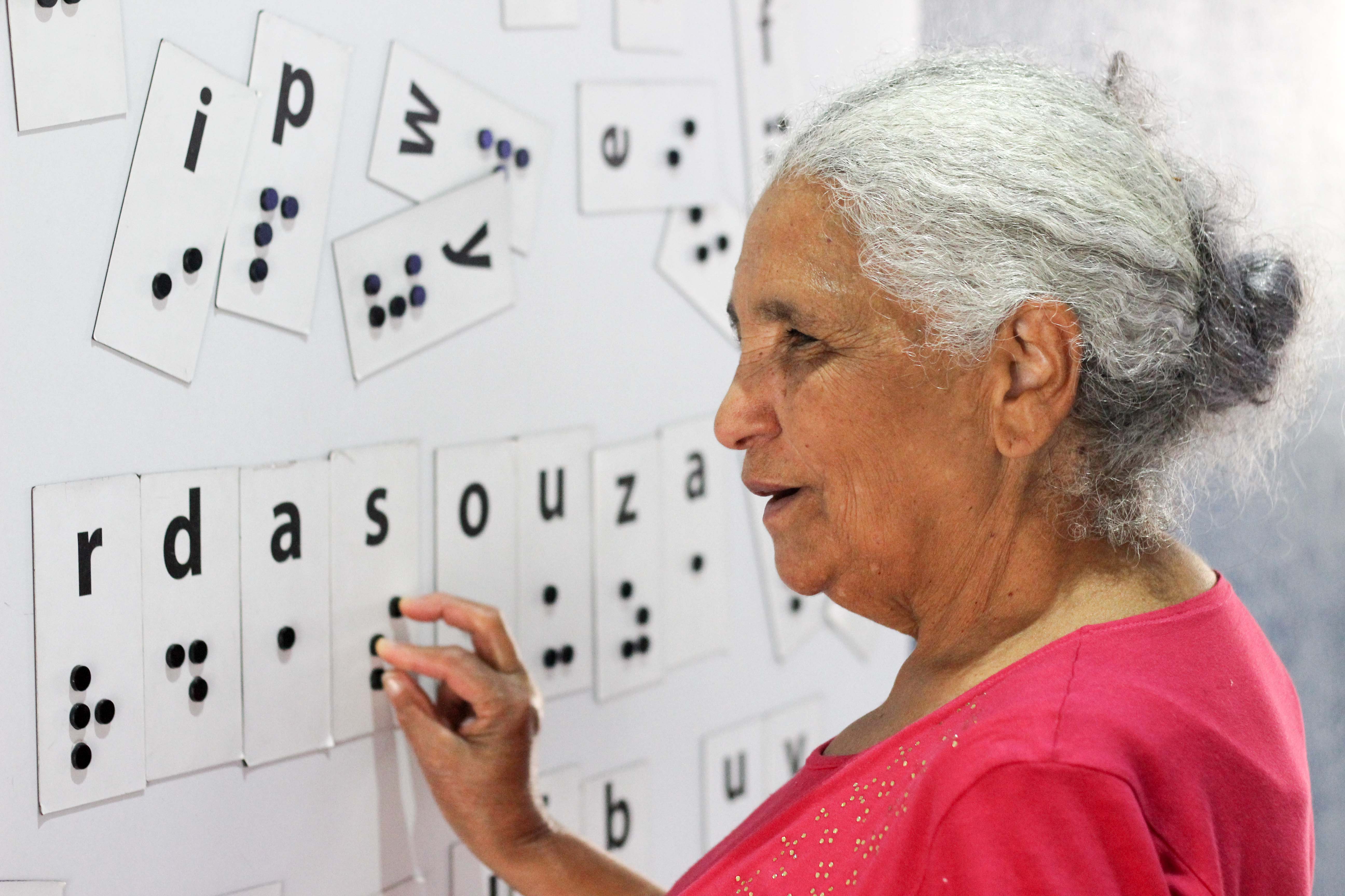 Descrição da imagem: foto de Dona Maria tateando um painel magnético com várias letras em braille embaralhadas. Ela está de perfil.