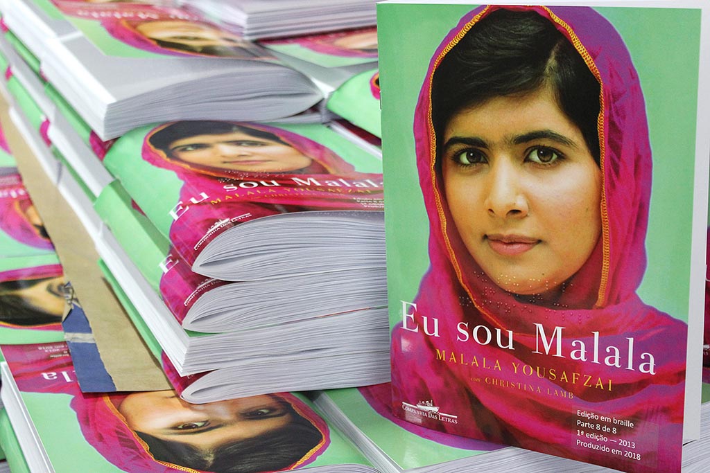 Descrição da imagem: foto do livro "Eu sou Malala" em uma pilha de outros exemplares do mesmo título. A capa traz a foto de Malala sorrindo  e usando um lenço rosa sobre fundo verde.