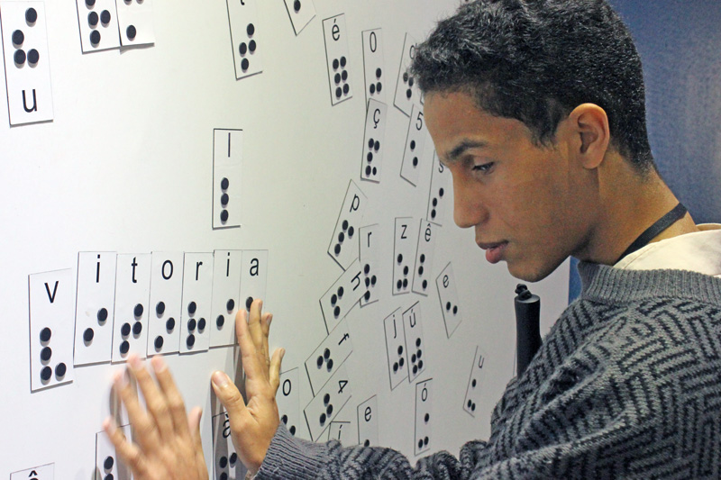 Descrição da imagem: foto de Lucas tocando um painel magnético com diversas letras em braille. Ele está de perfil, com as duas mãos sobre a palavra "vitória" afixada no painel.