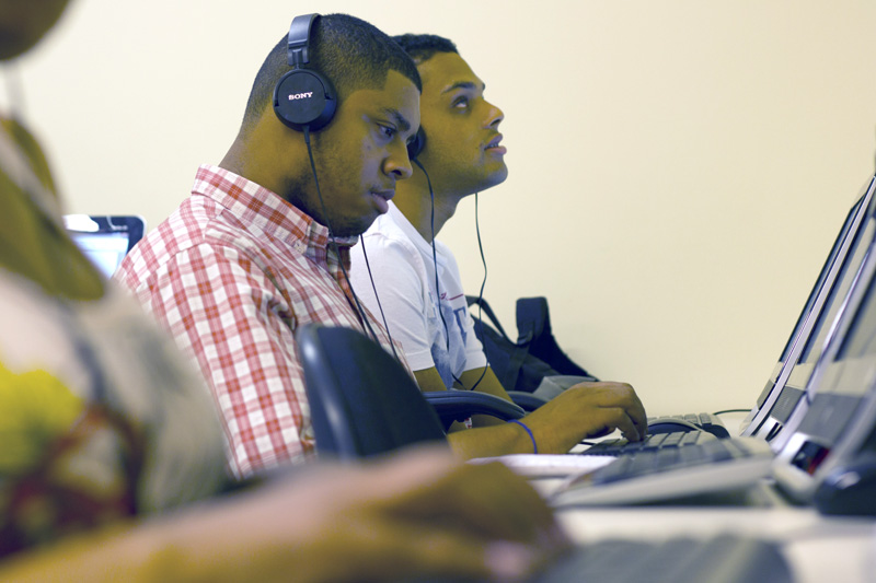 Descrição da imagem: foto de dois rapazes de aproximadamente 22 anos à frente de computadores. Eles estão de perfil e usam fones de ouvido. Na parte inferior da imagem é possível ver a mão de uma terceira pessoa sobre o teclado, na mesma mesa de trabalho.