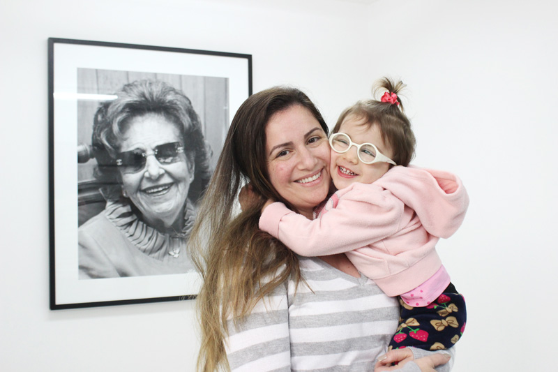 Descrição da imagem: foto de Nadia com Lívia no colo. A menina dá um abraço apertado na mãe. As duas olham pra frente e sorriem. Ao fundo há um retrato em preto e branco de Dorina Nowill na parede. 
