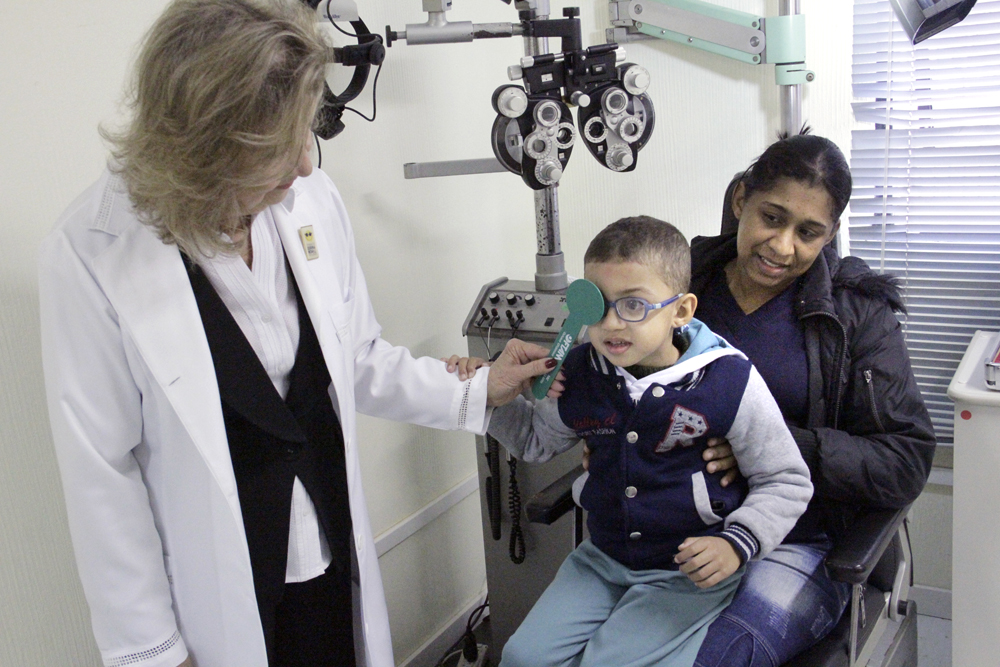 Descrição da imagem: foto de Marquinhos fazendo um exame oftalmológico. Ele está sentado no colo da mãe e segura o braço de uma profissional da Fundação Dorina, que o examina. Ela está de pé, cobrindo o olho direito de Marcos com uma haste de plástico verde. 