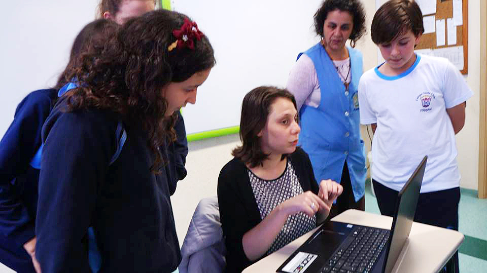 Descrição da imagem: foto de Isabela sentada em uma mesa escolar com um notebook à sua frente. Ao redor, de pé, estão três meninas, um menino e uma mulher de avental azul, olhando para o notebook.