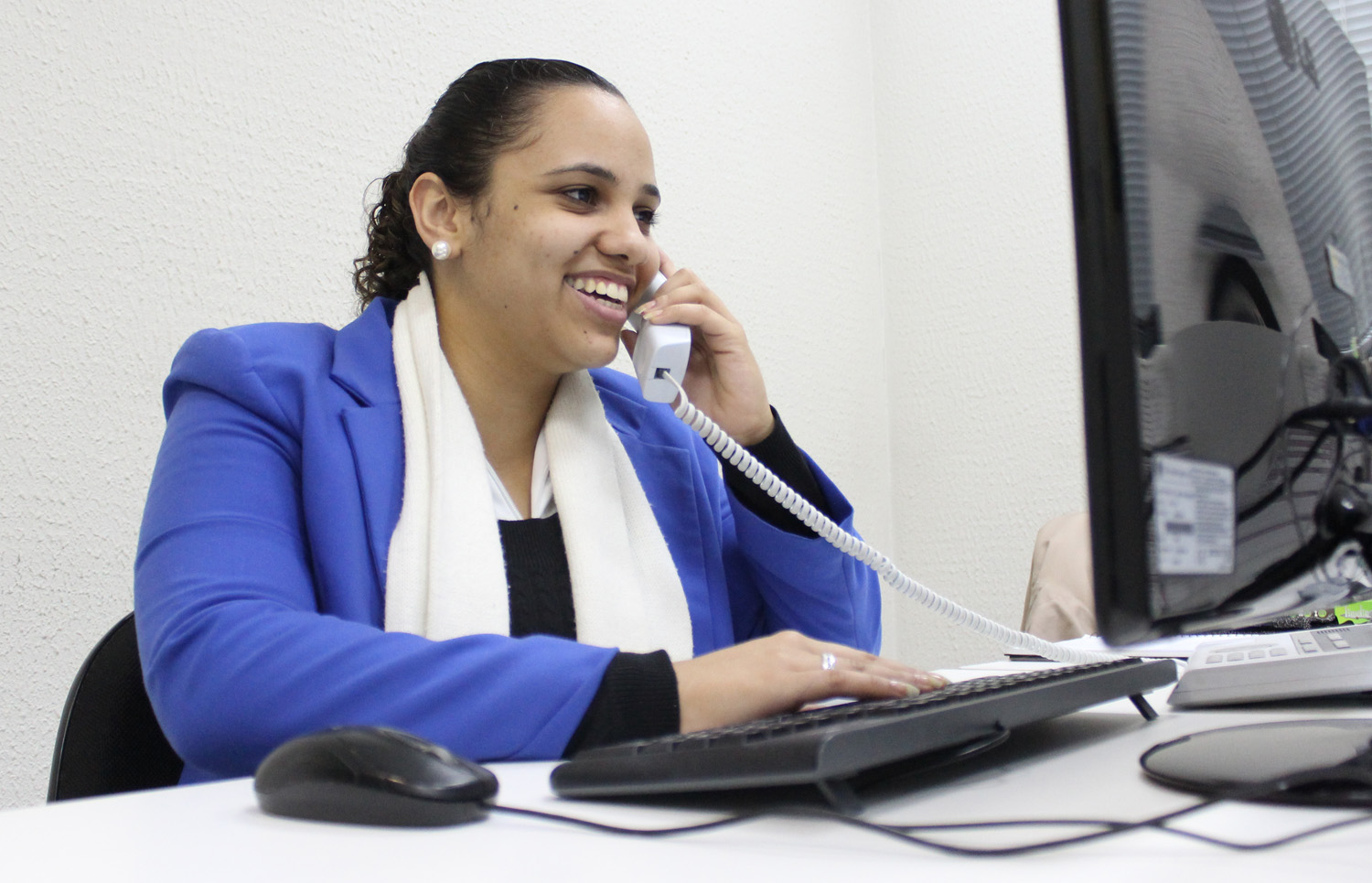 Descrição da imagem: foto de Stephani falando ao telefone numa mesa com computador. Ela está em meio perfil, olha em direção à tela e sorri.