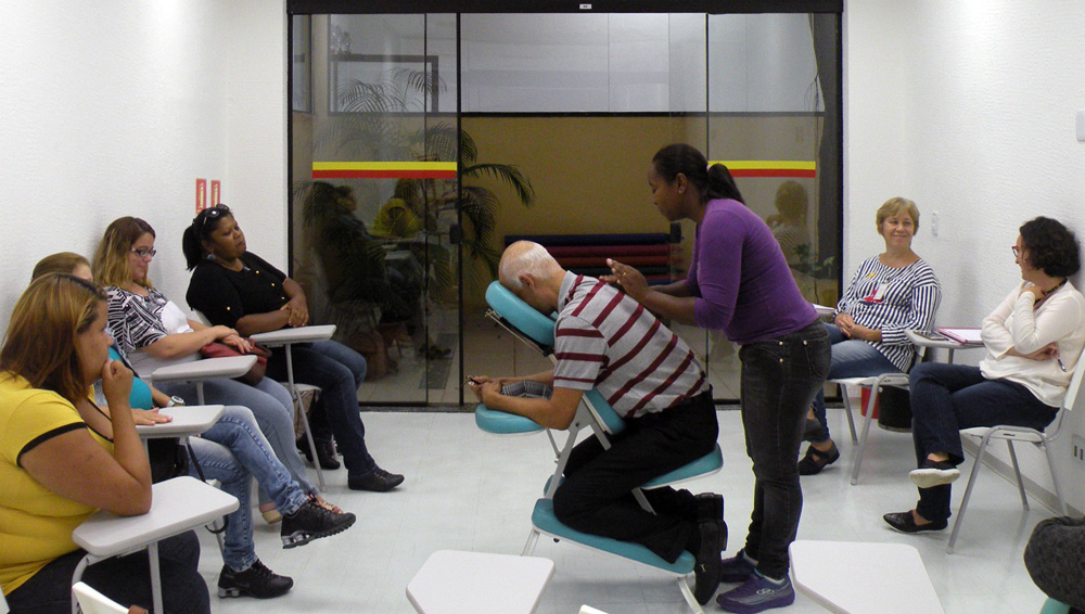 Descrição da imagem: foto de uma mulher fazendo massagem nas costas de um senhor, que está sentado numa cadeira de massoterapia em uma sala de paredes brancas e porta de vidro. Ao redor deles, sete pessoas sentadas em cadeiras universitárias observam.