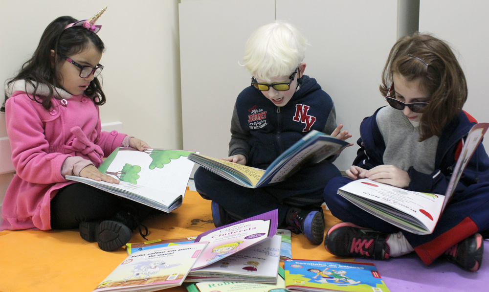 Descrição da imagem: foto de três crianças lendo livros. Todas estão sentadas no chão e usam óculos. 