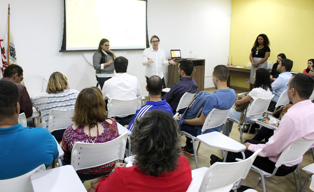 Descrição da imagem: foto de uma sala com dezenas de pessoas sentadas em cadeiras universitárias. À frente delas, de pé, há uma mulher e um homem que gesticula. Atrás deles, um telão em branco.