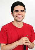 Descrição da imagem: foto de Lucas Borba retratado da cintura pra cima. Ele tem pele clara, cabelos pretos e curtos, usa camiseta vermelha, apoia as duas mãos sobre uma bengala e sorri.