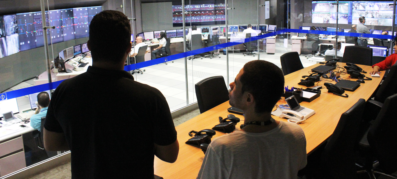 Descrição da imagem: foto de Lucas e um funcionário do metrô no Centro de Controle Operacional do Metrô. Eles estão de costas, olhando, através de uma parede de vidro, para uma sala repleta de painéis eletrônicos e computadores operados por cerca de 10 pessoas.