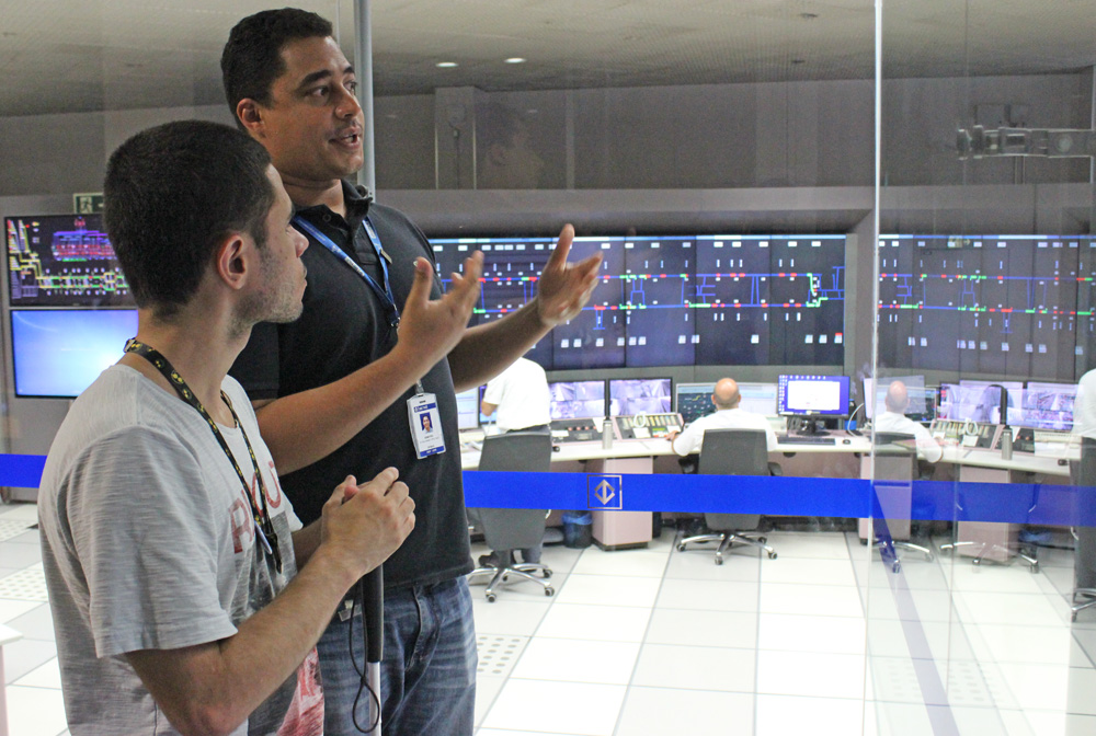 Descrição da imagem: foto de um funcionário do metrô conversando com Lucas no Centro de Controle Operacional. Ao fundo há painéis eletrônicos na parede e pessoas no computador.