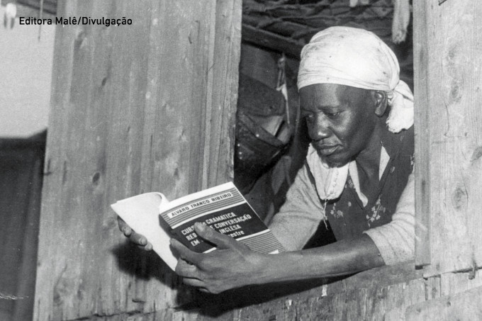 Descrição da imagem: foto em preto e branco de Carolina de Jesus lendo um livro. Ela está debruçada na janela de uma parede de madeira e usa um lenço branco na cabeça.