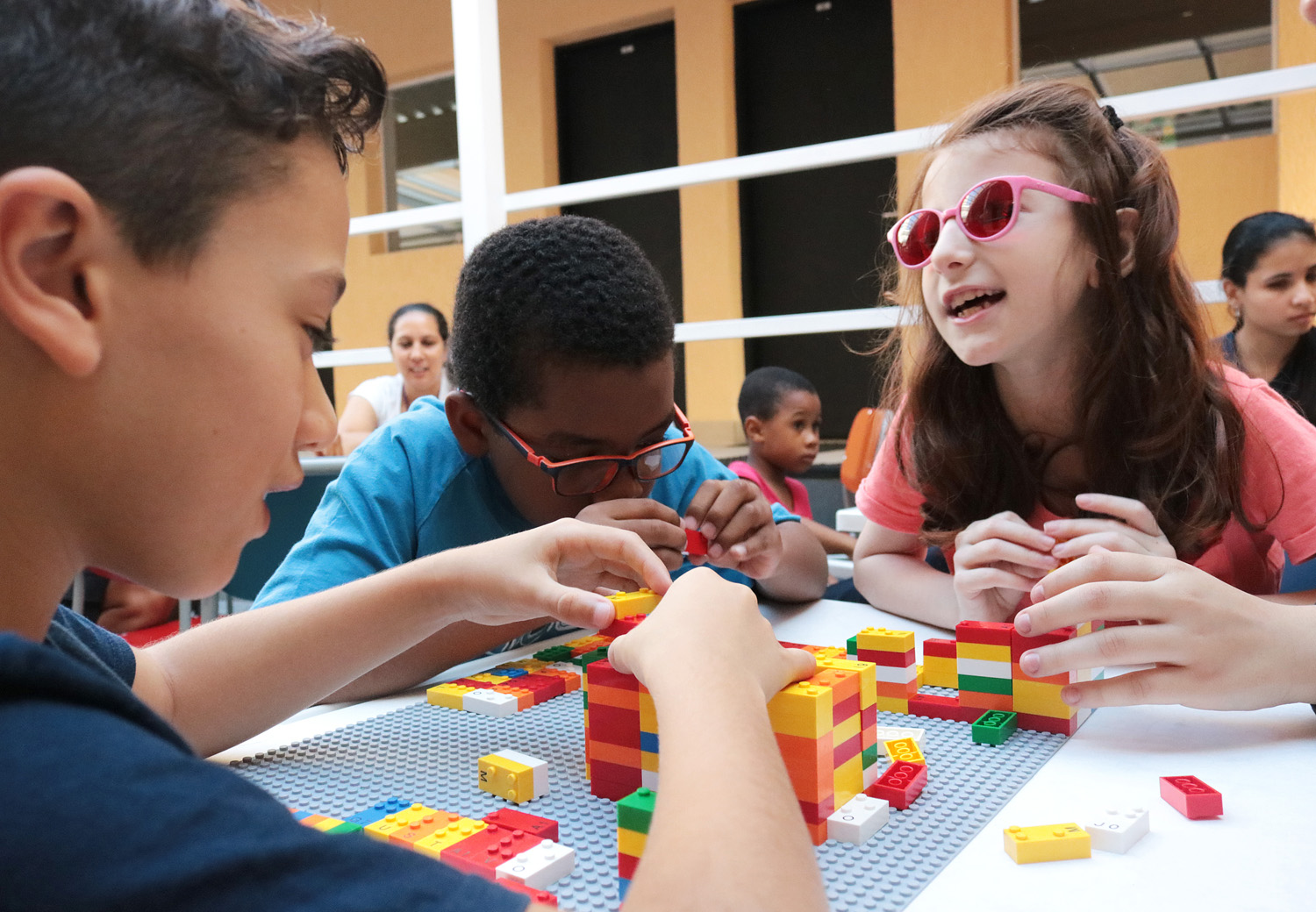 Descrição da imagem: foto de três crianças em volta de uma mesa manuseando peças do LEGO Braille Bricks. Ao fundo há duas mulheres e um menino.