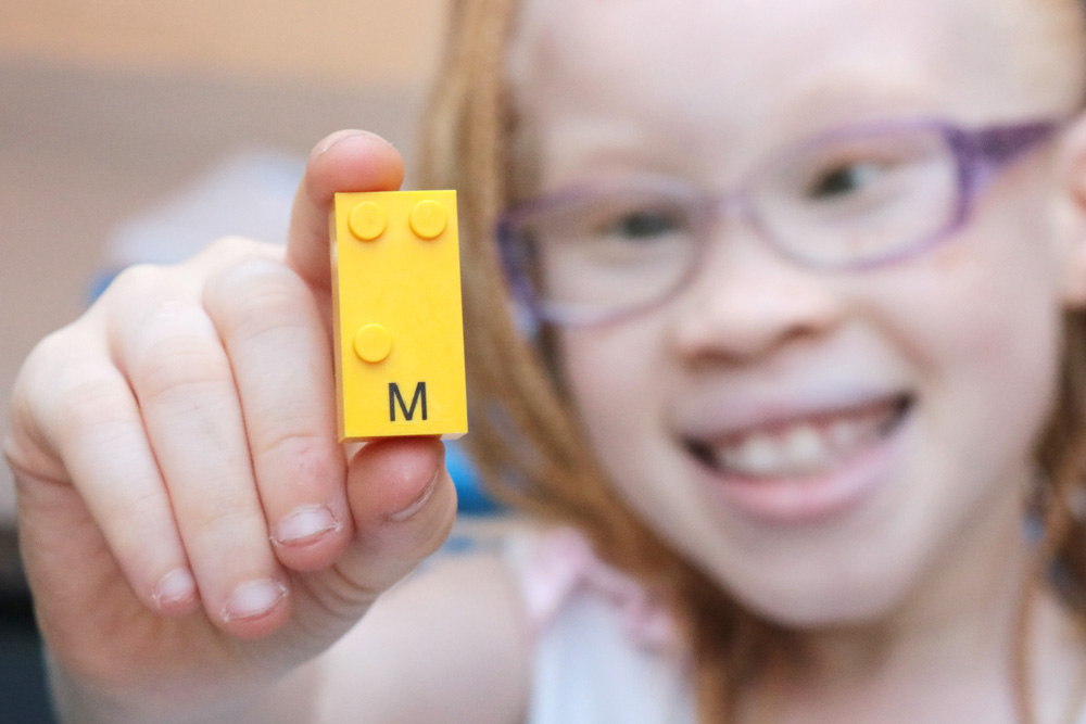 Descrição da imagem: Monica segura peça do Lego Braille Bricks. Ela está segurando a peça de letra M de cor amarela. Ela aparece ao fundo levemente desfocada enquanto mostra a peça para a câmera.